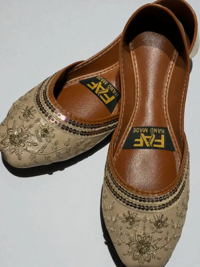 Pair of beige Punjabi Jutti flats with all-over gold zari thread and sequin embroidery, featuring a tan leather inner sole with a 'HAND MADE' label.