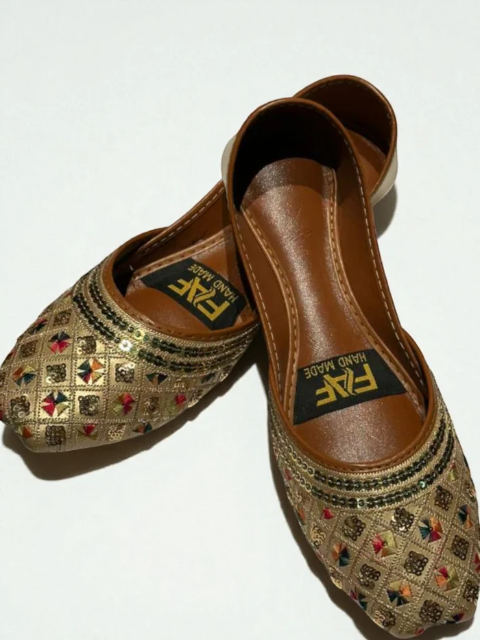 Pair of gold Punjabi Jutti flats with all-over geometric multi-colored zari thread and sequin embroidery, featuring a tan leather inner sole with a 'HAND MADE' label.