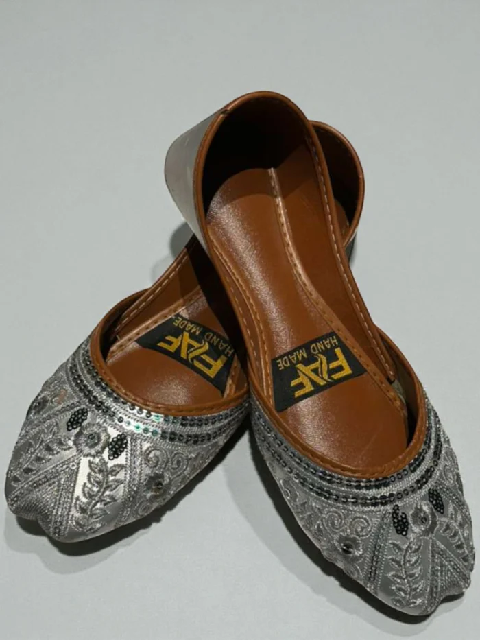 Pair of silver Punjabi Jutti flats with all-over silver zari thread and sequin embroidery, featuring a tan leather inner sole with a 'HAND MADE' label.