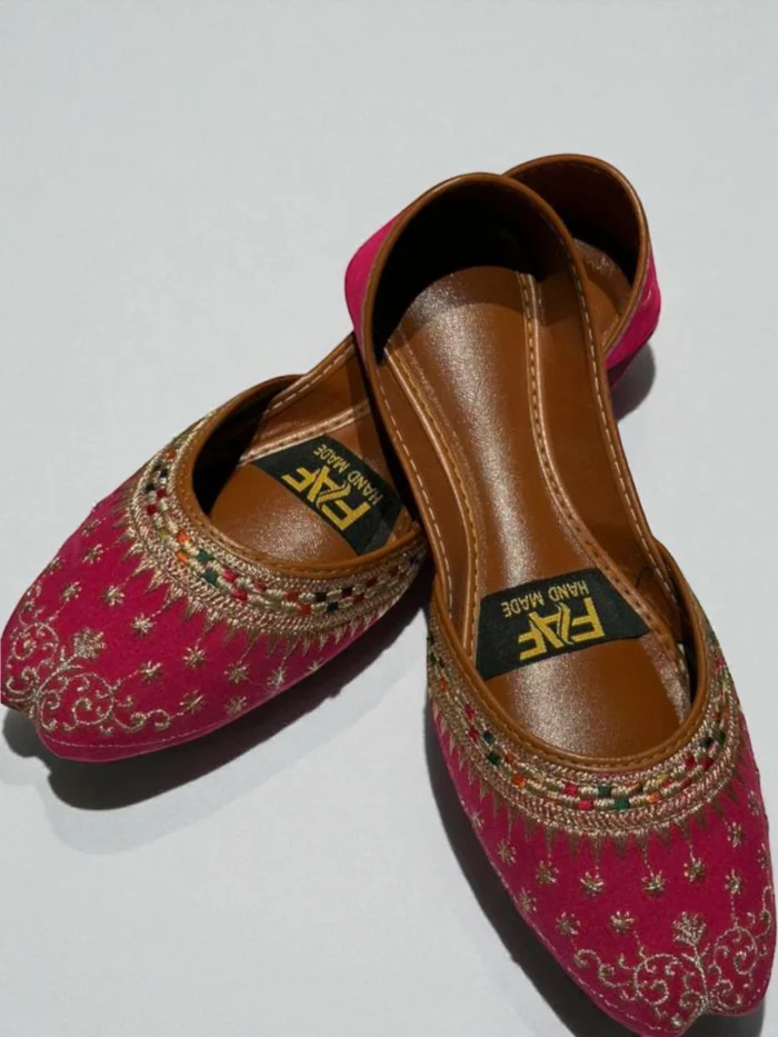 Pair of bright hot pink Punjabi Jutti flats with all-over gold zari thread and multi-color embroidery, featuring a tan leather inner sole with a 'HAND MADE' label.