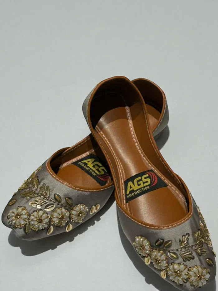 Pair of soft grey Punjabi Jutti flats with gold zari thread and white bead floral hand work, featuring a tan leather inner sole with an 'AGS Production' label.