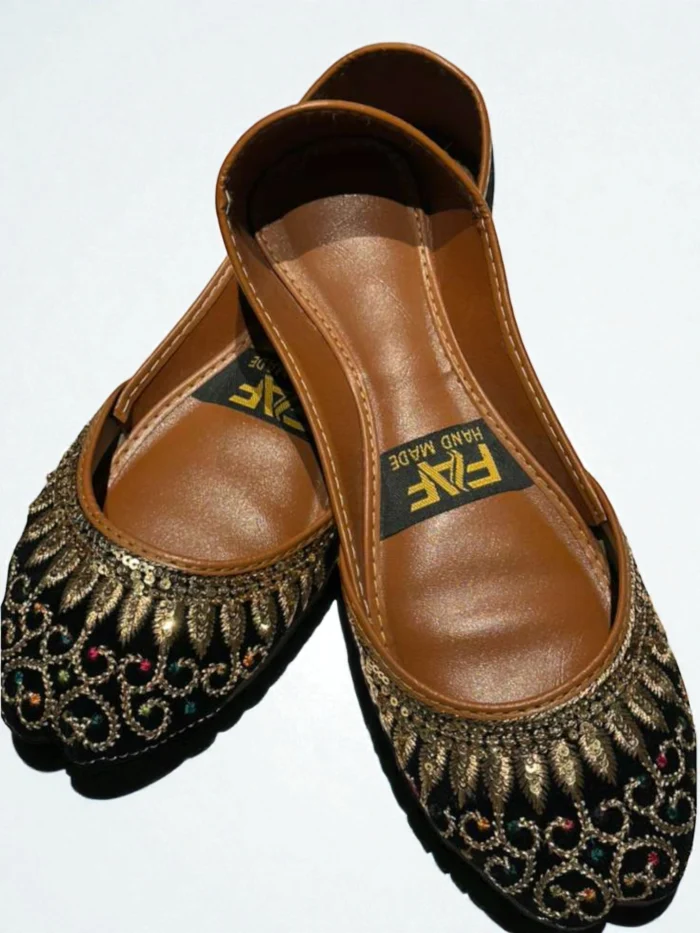 Pair of black Punjabi Jutti flats with all-over intricate gold zari thread and multi-color sequin work, featuring a tan leather inner sole with a 'HAND MADE' label.