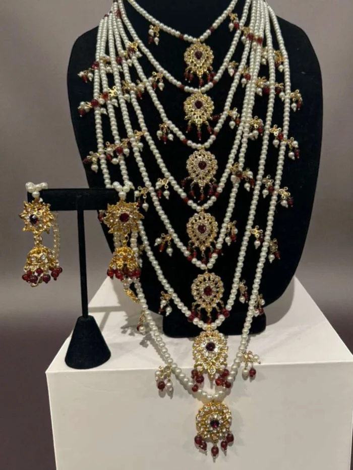 Magnificent gold plated 7-layer rani haar necklace with white faux pearls and red stone pendants, displayed on a black jewelry bust, with a matching earring set.