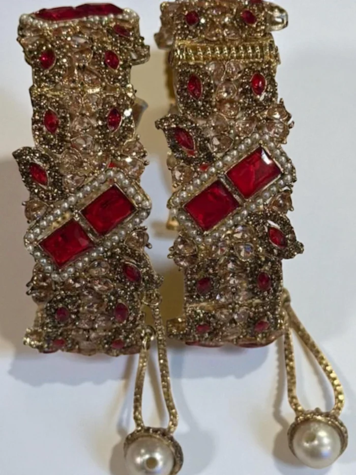 Pair of broad, openable gold plated kada bangles featuring stunning red rectangular and teardrop stones, clear accent stones, and a hanging pearl drop.