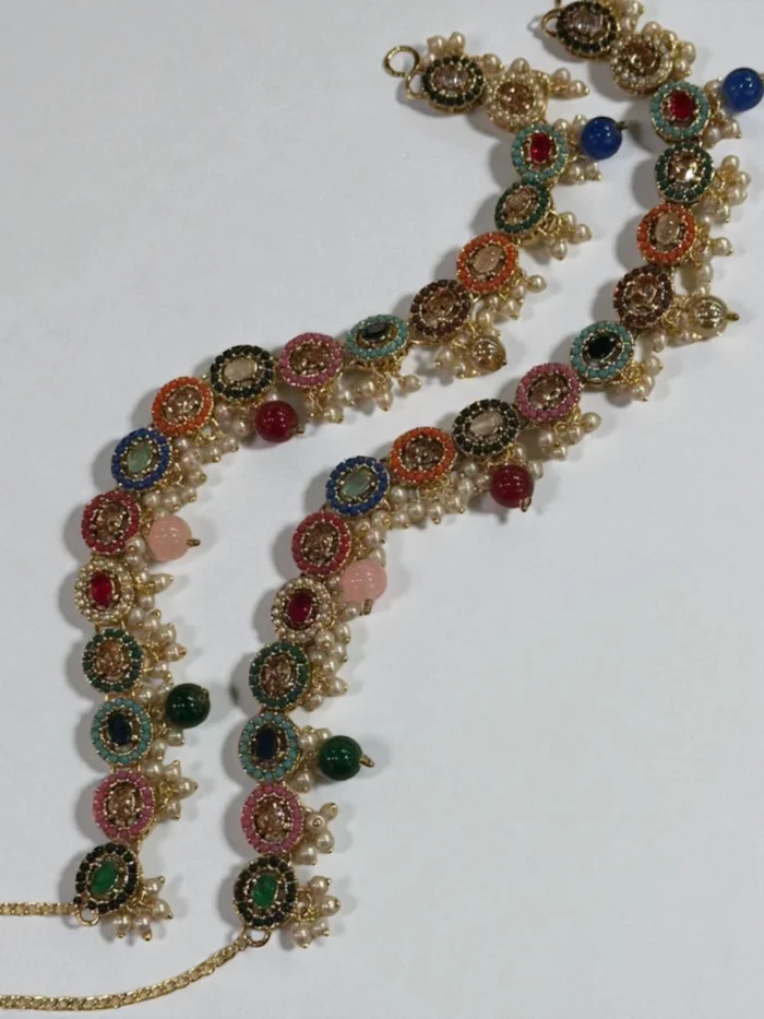 Pair of gold plated Nauratan anklets with linked round multi-colored stones and dangling faux pearl drops, shown curved on a white surface.